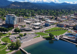 Aerial view of downtown Coeur d'Alene, Idaho, showcasing real estate opportunities with green park spaces, a parking lot, and nearby buildings. Surrounding mountains, trees, and a clear blue sky complete this picturesque setting.