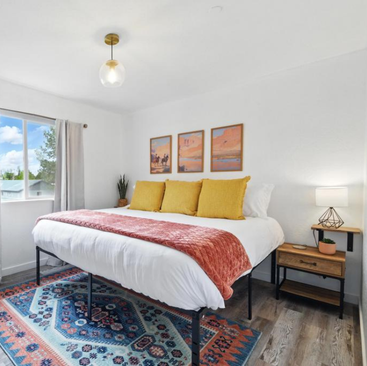 A bright Sandpoint vacation rental bedroom with a large bed featuring yellow pillows and a pink throw, a patterned rug, bedside table with lamp, and three framed art pieces. Sunlight streams in through a window with gray curtains.
