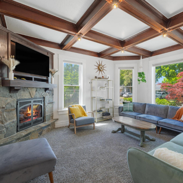 Bright living room with large windows, a stone fireplace, wood ceiling beams, and gray furniture. Green plants and colorful pillows add charm. Sunlight fills this inviting CDA Vacation Rental in beautiful Coeur dAlene.