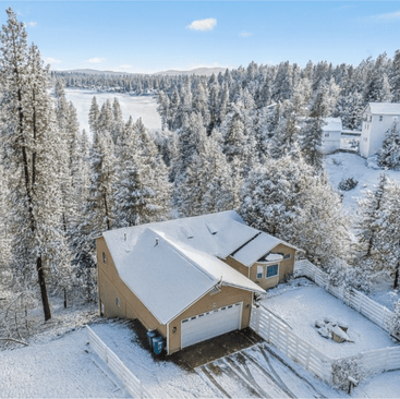 A tan house with a snow-covered roof and driveway sits near Hayden Lake, surrounded by tall, snow-dusted pine trees and a white fence, set in a forested winter landscape under a bright blue sky.