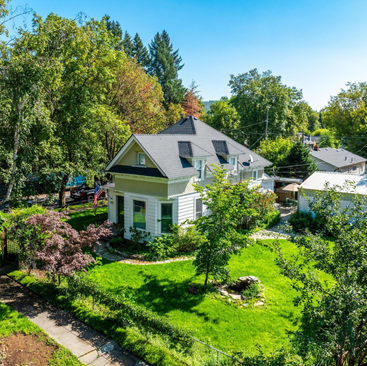 Aerial view of a charming white house with a dark roof, surrounded by lush green grass, trees, and a stone walkway—perfectly located near midtown, ideal as your next CDA vacation home in a sunny suburban neighborhood.