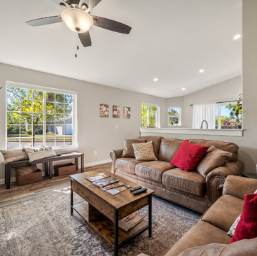 Bright living room with a ceiling fan, brown sofas with red and patterned cushions, a wooden coffee table, and large windows—perfect for relaxing in Idaho Vacation Rentals.