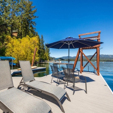1 Two lounge chairs and a table with four chairs sit on a wooden dock by a lake, shaded by a blue umbrella. Perfect for relaxing, this scene showcases the tranquility North Idaho Vacation Rental Management can offer under clear blue skies.