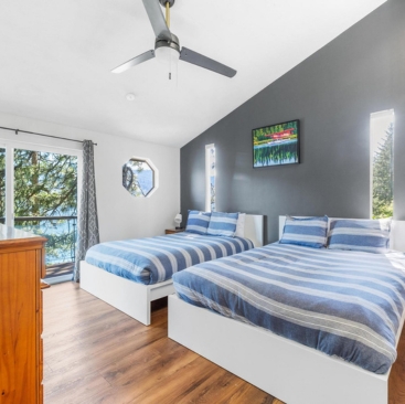 10 Bright bedroom with two double beds featuring blue striped bedding, a dark accent wall, wood flooring, ceiling fan, dresser, and large windows overlooking trees and a lake—professionally maintained by North Idaho Vacation Rental Management.