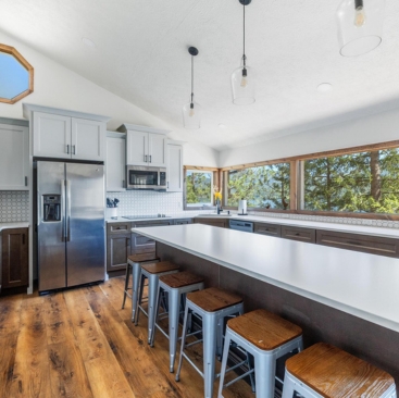 11 Modern kitchen with stainless steel appliances, gray cabinets, a large island with four stools, wood flooring, pendant lights, and large windows—perfect for enjoying trees and natural light in your North Idaho vacation rental management property.