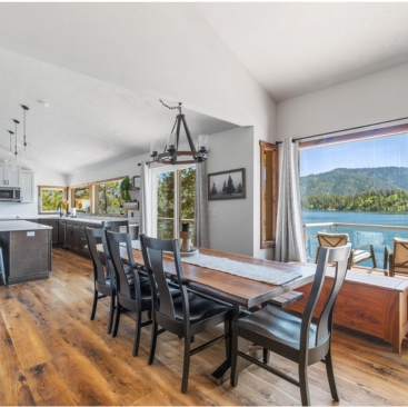 12 Spacious dining area with a wooden table and black chairs, open to a modern kitchen. Large windows offer scenic lake and mountain views, filling the room with natural light—perfectly managed by North Idaho Vacation Rental Management.