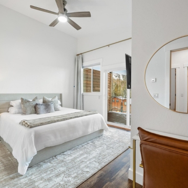 2 A modern bedroom, perfect for a North Idaho Vacation Rental Management listing, with a large bed, gray and white bedding, a rug, ceiling fan, round mirror above a brown chair, and glass doors opening to a balcony overlooking trees.