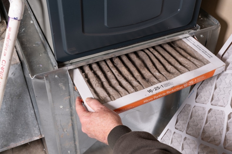 A person removes a dirty, pleated air filter from a furnace in North Idaho, revealing heavy dust buildup. Property owners or those focused on STR maintenance have a clean replacement filter ready to install for fresh indoor air.