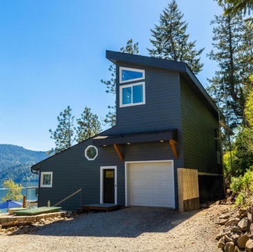 2 Modern dark blue house with large windows and an angled roof, managed by a professional Vacation Rental Property Manager, surrounded by tall trees and mountains, a gravel driveway, and a lake view in the background on a sunny day.