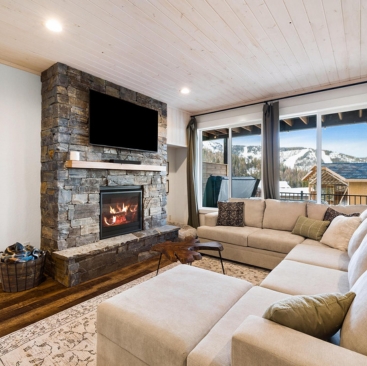 3 A cozy living room with a stone fireplace, wall-mounted TV, large beige sectional sofa, and windows displaying snowy mountain views—perfect for guests seeking North Idaho Vacation Rental Management in an inviting, warm space.