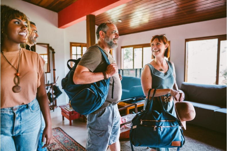 Four smiling adults with travel bags enter a bright, cozy living room with a wooden ceiling and large windows, reflecting the care taken during an Airbnb pre-arrival inspection to ensure everything is perfect for their stay.