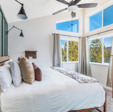 3 Bright bedroom with white bedding, assorted throw pillows, wall-mounted reading lights, and large windows with beige curtains letting in natural light and views of trees outside—perfectly managed by North Idaho Vacation Rental Management. Ceiling fan above the bed.