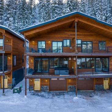 4 A large wooden cabin with two stories, balconies, and outdoor seating, surrounded by snow and pine trees. Warm lights glow inside and out, creating a cozy winter atmosphere—ideal for North Idaho Vacation Rental Management services.