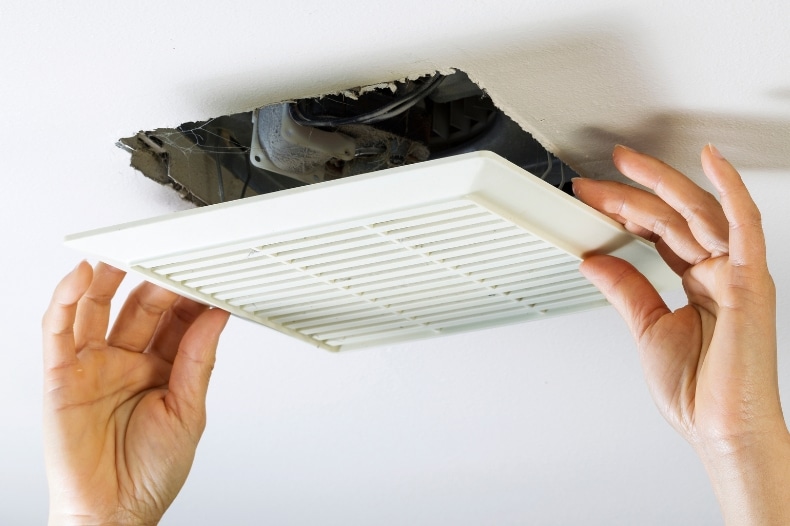 A person’s hands are installing or removing a white vent cover from a ceiling, revealing an opening with visible wires and ductwork inside—an important STR maintenance task for property owners in North Idaho.