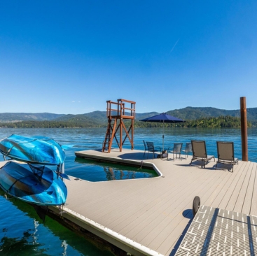 4 A wooden dock on a lake with lounge chairs, blue umbrella, stacked kayaks, and a diving platform—managed by a North Idaho Vacation Rental Management team—nestled amid mountains and forest under a clear blue sky.