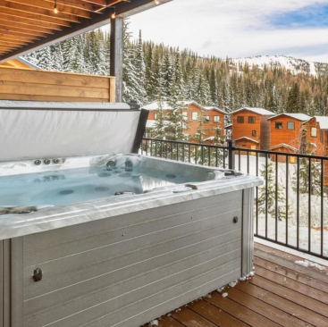 5 A hot tub sits on a wooden deck overlooking snow-covered trees and wooden cabins, managed by North Idaho Vacation Rental Management, with mountains in the background under a cloudy sky.