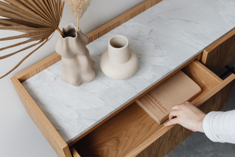A hand opens a wooden drawer beneath a marble-topped console table with two modern ceramic vases and dried plants—part of an Airbnb pre-arrival inspection. Inside the drawer is a closed brown notebook.