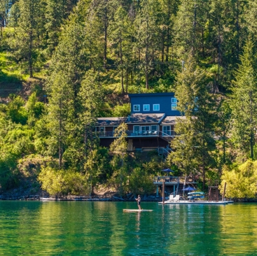 5 A person paddleboards on a calm lake in front of a house managed by a North Idaho Vacation Rental Management company, nestled among tall trees on a hillside with a dock stretching into the water.