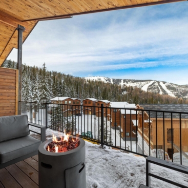 6 A cozy outdoor patio with a fire pit and gray seating overlooks snow-covered cabins and pine trees, with mountains beyond—a perfect retreat managed by North Idaho Vacation Rental Management.