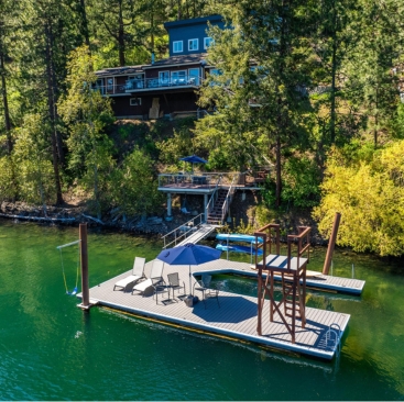 6 A lakeside home surrounded by trees overlooks a dock with lounge chairs, an umbrella, and a swimming platform on clear green water. Multiple decks connect the house to the waterfront—perfect for North Idaho Vacation Rental Management.