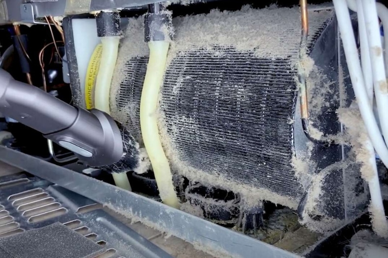 A vacuum cleaner nozzle is being used to remove a thick layer of dust from the condenser coils and surrounding parts at the back of a refrigerator—a helpful STR maintenance tip for North Idaho property owners. White wires and yellow tubes are also visible.