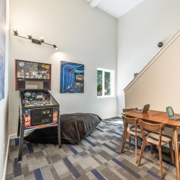 7 A modern room managed by a Vacation Rental Property Manager, featuring a pinball machine, bean bag, abstract wall art, small window, and a wooden desk with two chairs on a blue and gray patterned carpet.