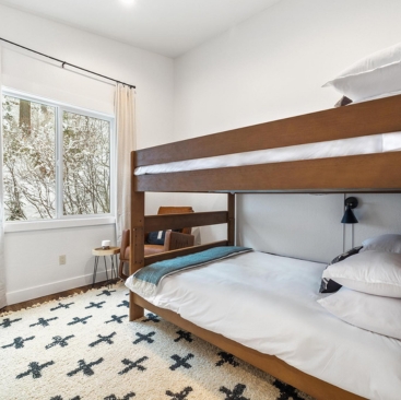 8 A bright bedroom with wooden bunk beds, white bedding, a patterned rug, and a cozy desk—perfect for guests seeking comfort. Managed by a North Idaho Vacation Rental Management team, with trees visible outside the window.