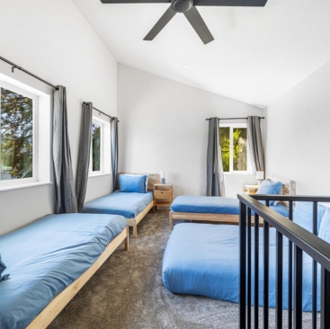 8 Bright bedroom with three single beds with blue bedding, grey curtains, large windows letting in natural light—a relaxing retreat managed by North Idaho Vacation Rental Management. Ceiling fan, nightstands, gray carpet, and black railing complete the space.