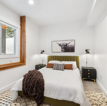 9 A cozy bedroom in a North Idaho Vacation Rental Management home features a green upholstered bed, white bedding, rust pillows, black nightstands with sconces, a bull print above the bed, and a window with wooden trim on the left.