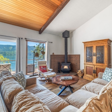 9 A cozy living room with a beige sectional sofa, patterned pillows, and a wood stove. Large windows open to a balcony overlooking a scenic lake—perfect for relaxing or seeking North Idaho Vacation Rental Management expertise.