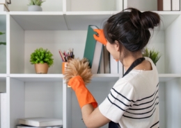 A woman wearing an apron and orange gloves is dusting shelves with a duster in a modern, white shelving unit—reflecting the pristine care provided by Haven Property Management for vacation rentals.