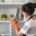 A woman wearing an apron and orange gloves is dusting shelves with a duster in a modern, white shelving unit—reflecting the pristine care provided by Haven Property Management for vacation rentals.