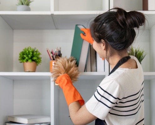A woman wearing an apron and orange gloves is dusting shelves with a duster in a modern, white shelving unit—reflecting the pristine care provided by Haven Property Management for vacation rentals.