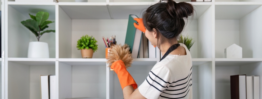 A woman wearing an apron and orange gloves is dusting shelves with a duster in a modern, white shelving unit—reflecting the pristine care provided by Haven Property Management for vacation rentals.