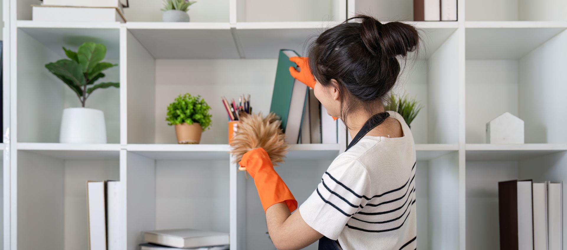 Why Every Airbnb Needs a Pre-Arrival Inspection System A woman wearing an apron and orange gloves is dusting shelves with a duster in a modern, white shelving unit—reflecting the pristine care provided by Haven Property Management for vacation rentals.