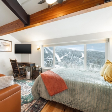Mogul Hill- Slideshow - 367x366 Cozy bedroom with large window overlooking snowy mountain landscape, bed with green quilt and orange throw, desk and TV, wooden beams, and warm lighting—perfect for guests seeking a North Idaho Vacation Rental Management experience.