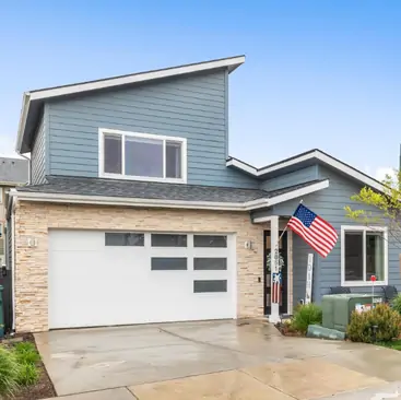 1 A modern two-story house with blue-gray siding, stone accents, a white garage door with glass panels—ideal for North Idaho Vacation Rental Management—features an American flag by the entrance and a wet driveway under a clear blue sky.