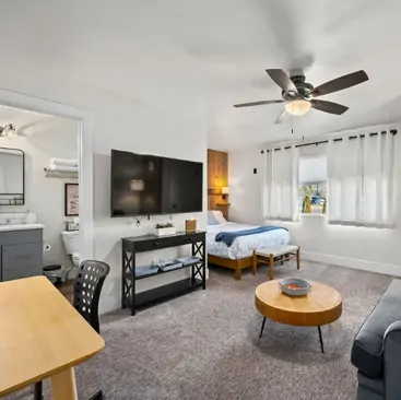 10 A cozy studio apartment managed by a Vacation Rental Property Manager, featuring a bed, TV, desk, small sofa, round coffee table, and a window with white curtains. The bright room is neatly organized with the bathroom visible through an open door.