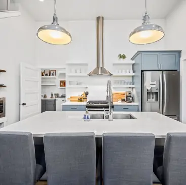 11 Modern kitchen with a large white island, four gray chairs, stainless steel appliances, blue cabinets, and open shelving—ideal for showcasing North Idaho Vacation Rental Management’s stylish properties. Two large pendant lights hang from the ceiling.