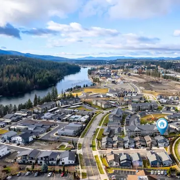 14 Aerial view of a suburban neighborhood near a river, with rows of houses, tree-lined streets, and forested hills in the background. A blue location pin highlights one home—an ideal spot for North Idaho Vacation Rental Management services. The sky is partly cloudy.