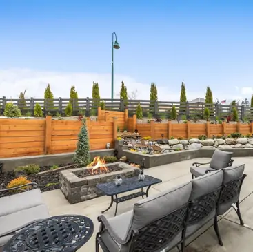 2 A modern patio with cushioned seating and a metal table surrounds a lit fire pit—ideal for gatherings. Managed by North Idaho Vacation Rental Management, the area features tiered landscaping, small trees, and a wooden fence under a bright blue sky.