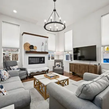 3 Modern living room with grey sofas and armchairs, a wooden coffee table, wall-mounted TV, fireplace, and large windows with roller shades. This inviting space is expertly styled by a top North Idaho Vacation Rental Management team.