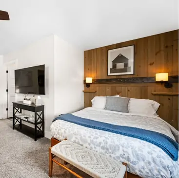 5 A cozy bedroom managed by North Idaho Vacation Rental Management features a large bed with a blue blanket, decorative pillows, and a wooden accent wall. Enjoy the barn photo, TV on a black console, and a patterned bench at the foot of the bed.