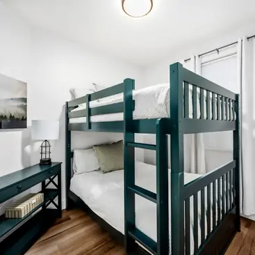 6 A small bedroom with teal bunk beds, white bedding, a black desk with books and a lamp, wood floor, white curtains, and a landscape painting. Natural light fills the space—perfect for those seeking North Idaho Vacation Rental Management comfort.
