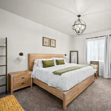 8 A cozy bedroom in this Vacation Rental Property Manager’s listing features a wooden bed, white bedding, green accents, a ladder shelf, modern chandelier, window with white curtains, and a chair by the floor mirror—perfect for your North Idaho getaway.