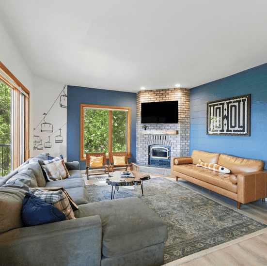 A bright living room in this Sandpoint vacation rental features a gray sectional, tan leather sofa, blue accent wall, brick fireplace with TV, large windows, and modern decor—perfect for relaxing after a day at Schweitzer ski resort in Idaho.
