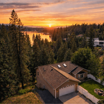 A tan house with a two-car garage sits among tall pine trees, overlooking Hayden Lakeview at sunset. The sky glows orange and purple as sunlight reflects on the water—a peaceful oasis perfect for Hayden Lake Vacation Rentals.