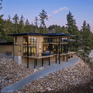 Modern two-story house with large glass windows and wood paneling, set on a rocky hillside near Lake Pend Oreille, surrounded by pine trees at sunset. A spacious deck runs along the front, offering scenic views—ideal for Sandpoint vacation rentals.