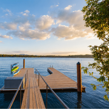 A wooden dock extends over a calm lake under a partly cloudy sky at sunset. Colorful kayaks are stacked on a rack to the left, and leafy branches frame the scene.