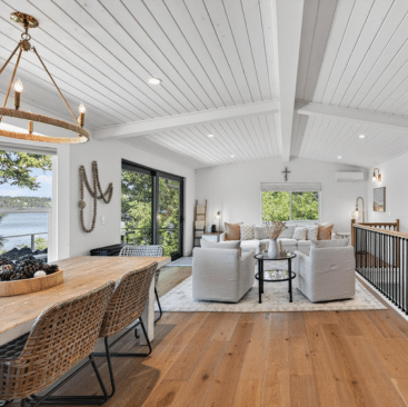 Bright, modern living and dining area with white walls and ceiling, wood floors, wicker dining chairs, cozy seating, large windows, and sliding doors overlooking a scenic outdoor view.